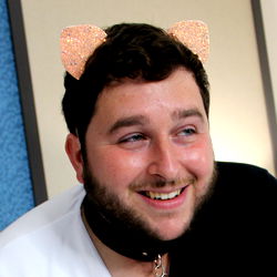 A model with a fun and creative appearance, wearing an animal-shaped ear accessory and a cat collar with sparkles. He's smiling in the photo and appears to be relaxed.