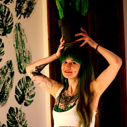 Lea, a model from Texas with a tattoo and green hair, standing in front of a plant wall holding a potted plant on her head.