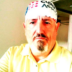 Shag69, a man with a beard, wearing a bandana and shirt, poses for a photo in his home.