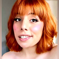 A young adult woman with short red hair, wearing a light pink lipstick and a white top with a subtle pink color near the collar. She is smiling subtly and has a glamorous style. Onlyfans model name: Madison Dakota