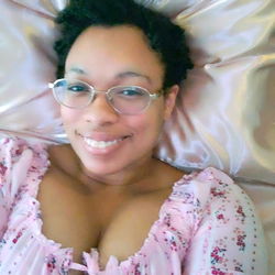 Amira Yums, a model with glasses and curly hair relaxing on a bed.