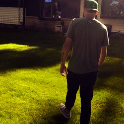 A young man in a green shirt, walking towards the camera through a backyard with a house in the background.