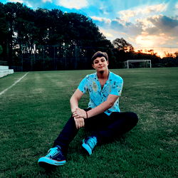 Model Pedro Eduardo, 19, poses in a soccer field with vibrant green grass and a clear sky.