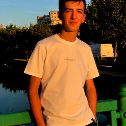 A young man posing in an urban setting, smiling and looking directly at the camera.