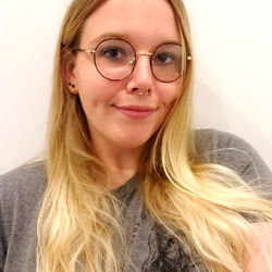 Miss Lily XO: A young woman with glasses and a ring piercing, posing against a white wall in a gray t-shirt.