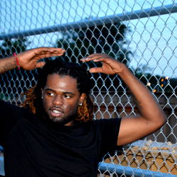 Shareef Papi, a rapper leaning on a fence with his hand on his head,