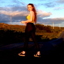 Lucy, a young woman walking down a country road, looking towards the camera with her hair in a ponytail. She's wearing casual athletic attire and black sneakers. The background shows an open field under a partly cloudy sky.