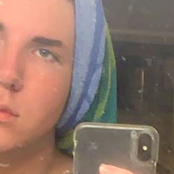 A young male, Drew Decker, with blue hair and a towel headscarf, captures his reflection in a mirror with an iPhone. He is looking directly at the camera with a thoughtful expression.
