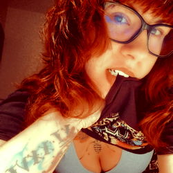 Red-haired OnlyFans model with glasses, showcasing her tattoos and a black t-shirt.