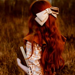 A captivating photograph of a model with red hair and freckles, set against a field of tall grass. The model is holding a small book and wears a plaid dress with a bow in their hair.